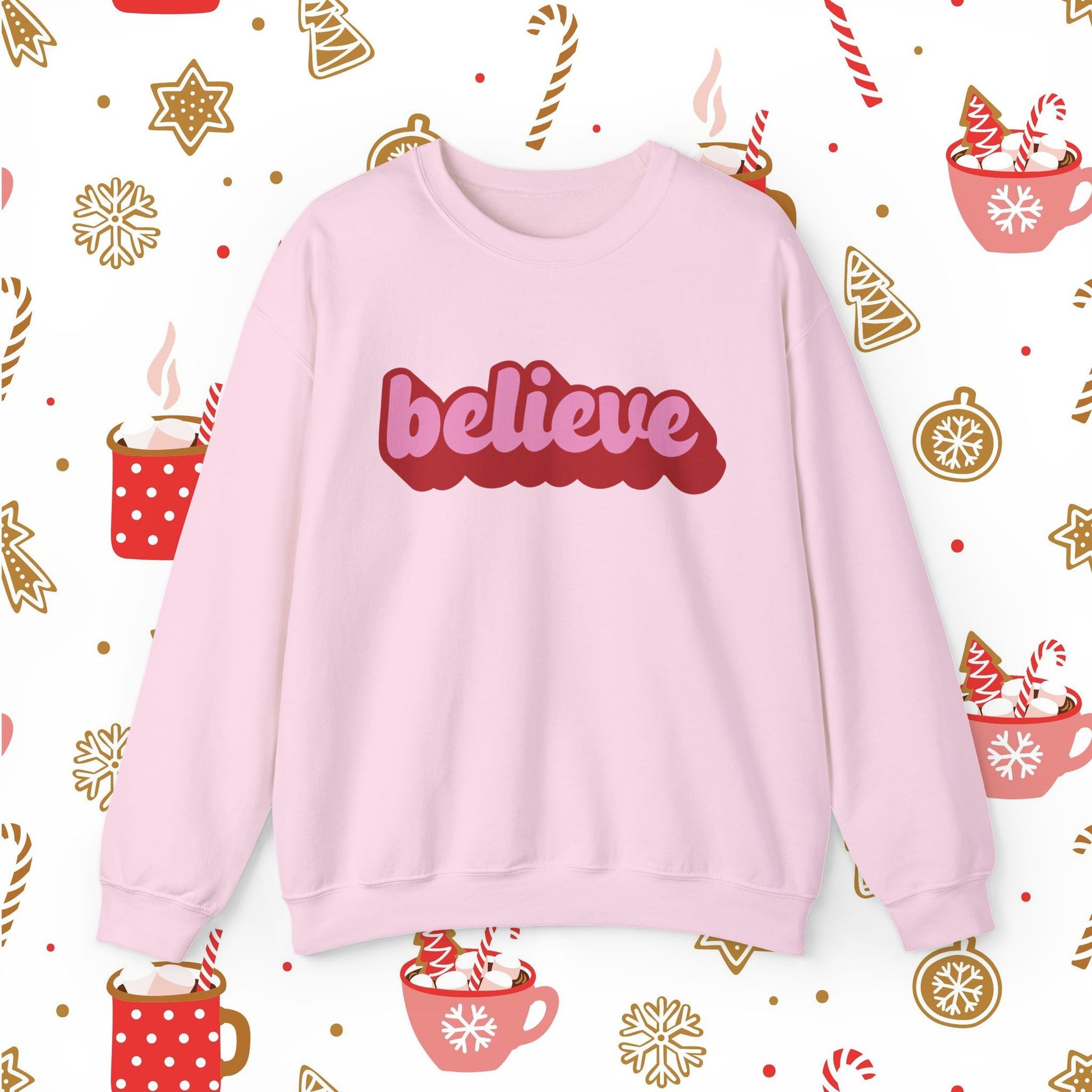 Pink sweatshirt with 'believe' text on a festive background with hot chocolate cups and candy canes.