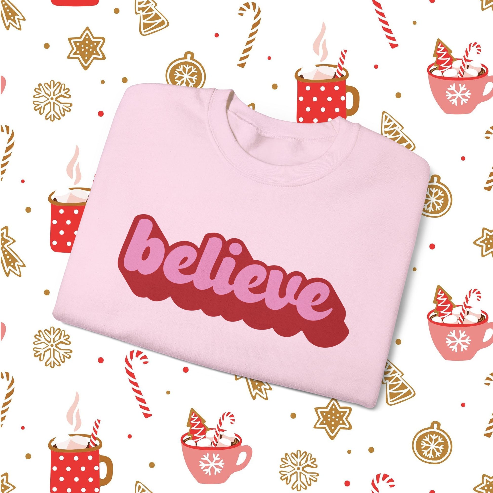 Pink sweatshirt with 'believe' text on a festive background with hot cocoa and candy cane illustrations.