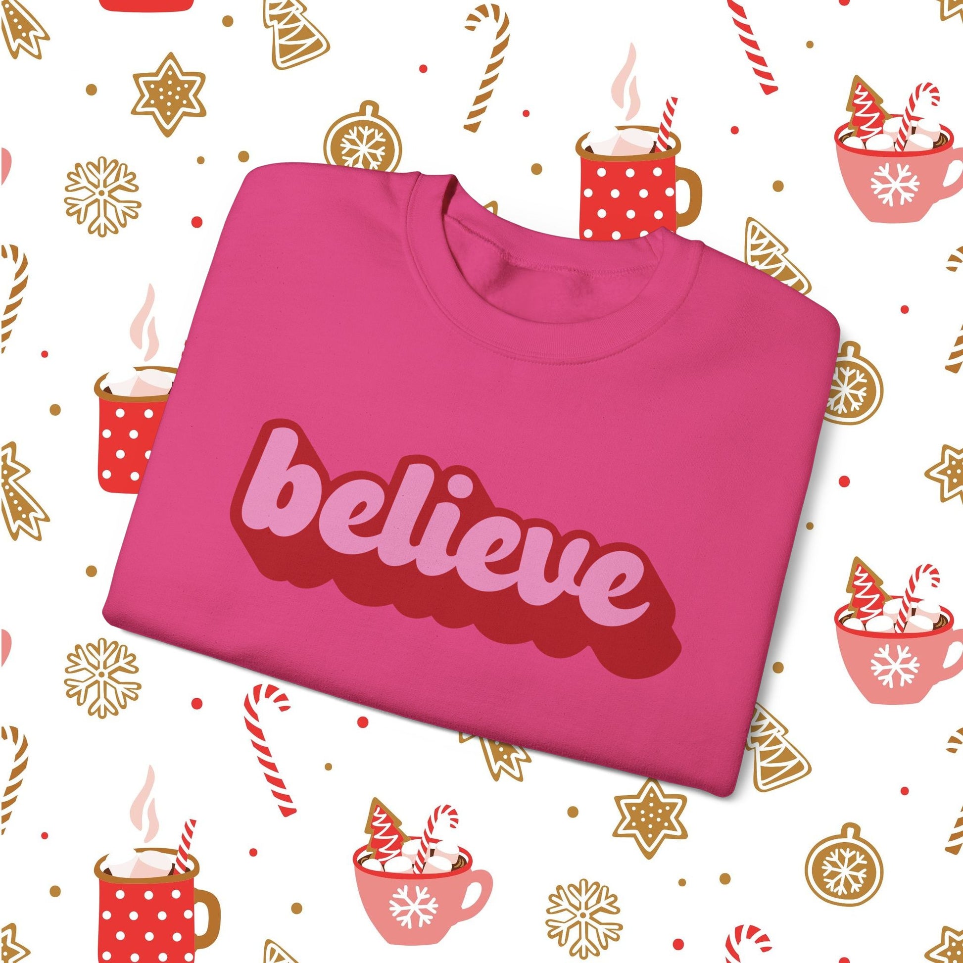 Pink sweatshirt with 'believe' text on a festive background with hot cocoa and candy cane illustrations.