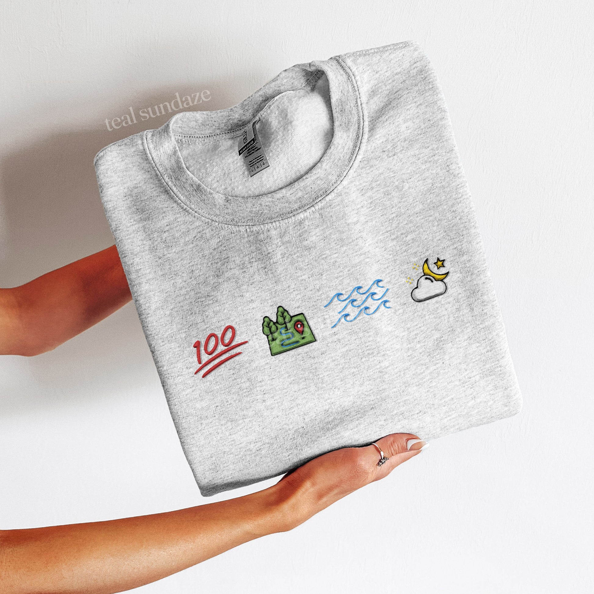 Folded gray sweatshirt with colorful ophelia themed embroidered icons representing 100, land, sea, sky held by a person on a light background