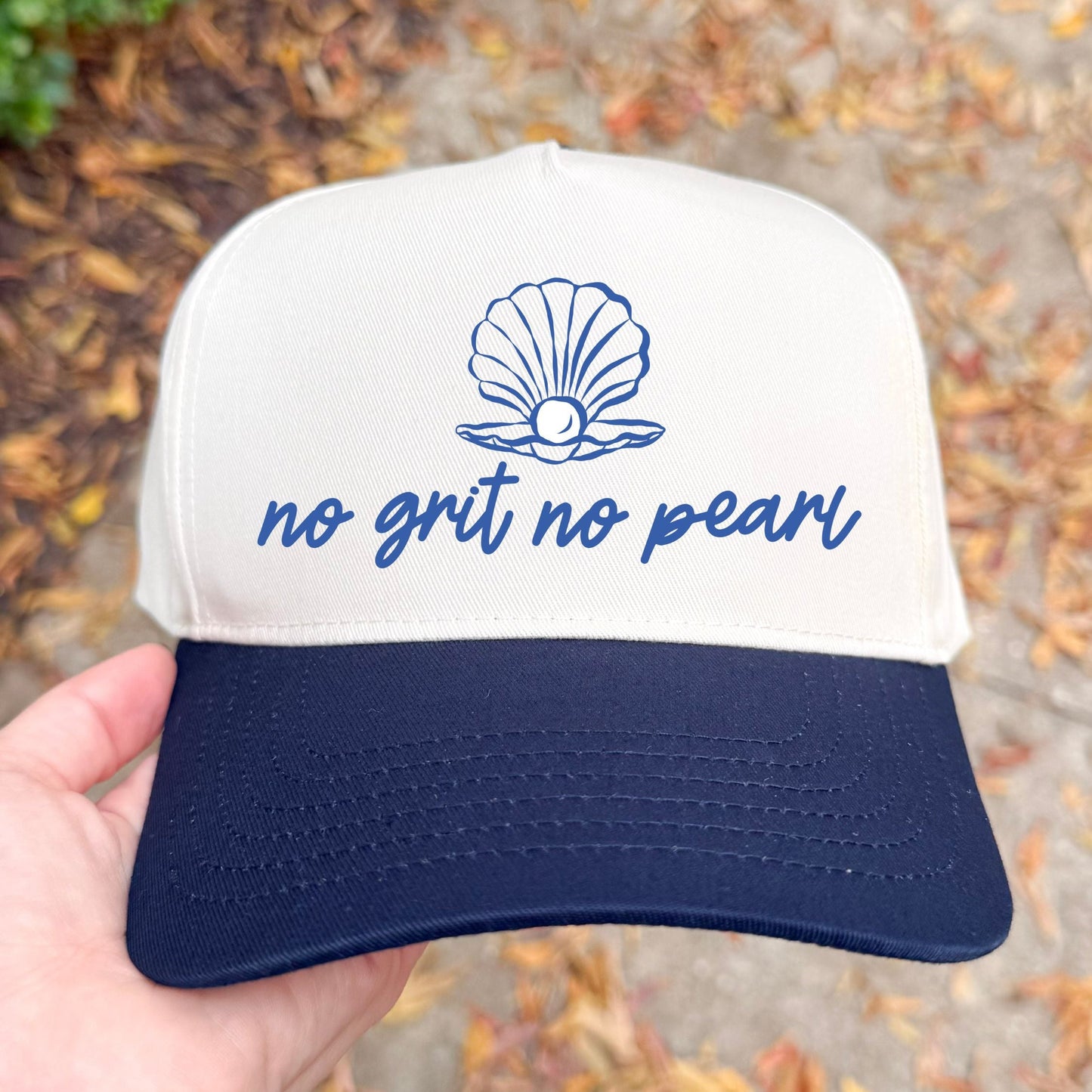 natural and navy cap with 'no grit no pearl' text and shell graphic held by a hand outdoors.