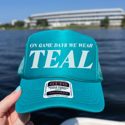 Turquoise cap with 'On game days we wear teal' text held over a body of water.