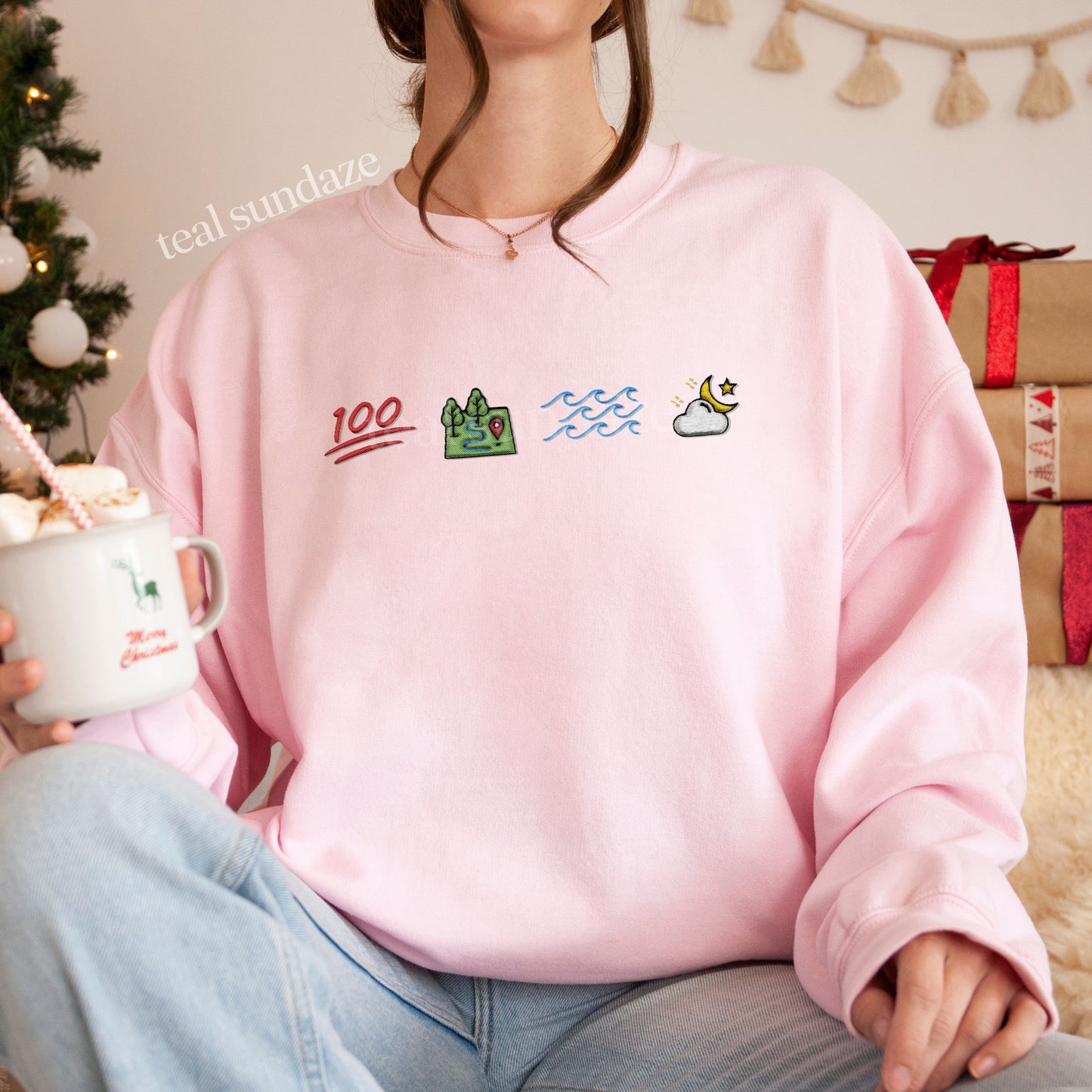 Person wearing a pink sweatshirt with Ophelia themed icons representing 100, land, sea sky, holding a mug in a cozy indoor setting.