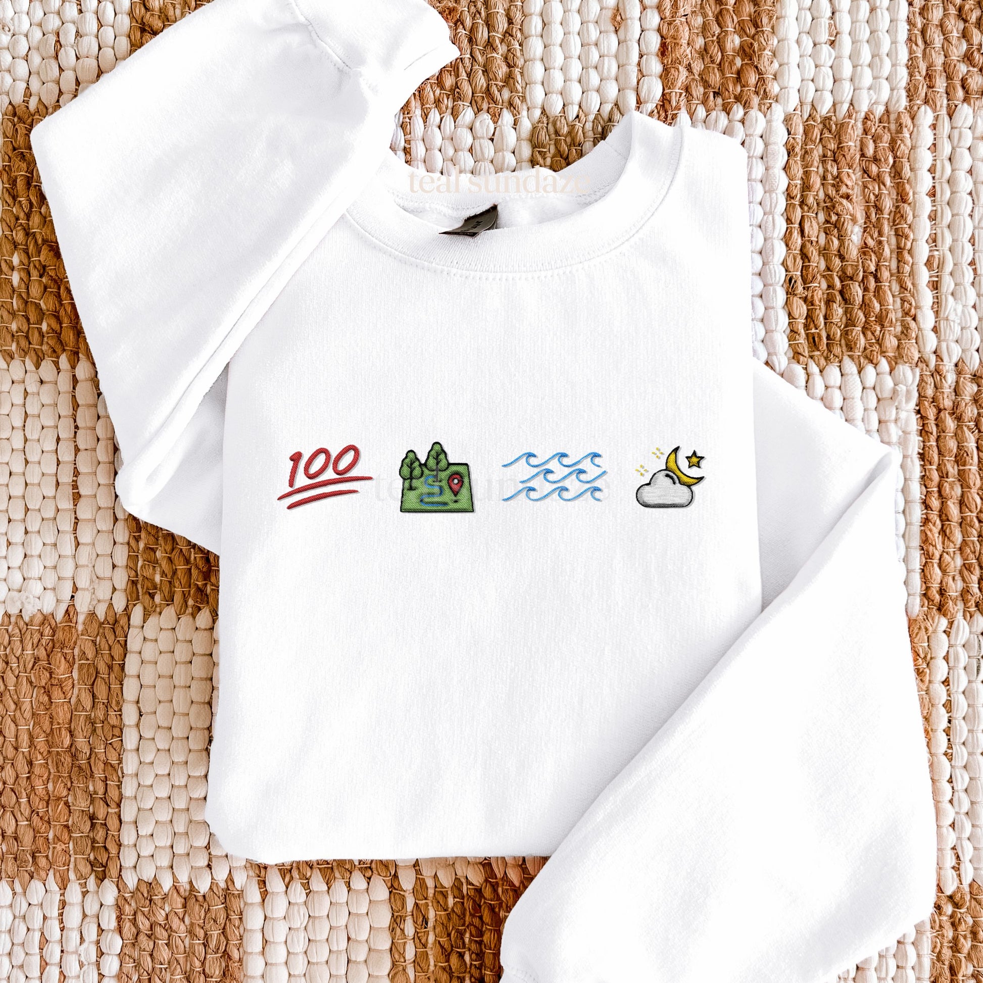 White sweatshirt with Opehlia themed embroidered icons representing 100, land, sea, sky on a textured brown and beige surface